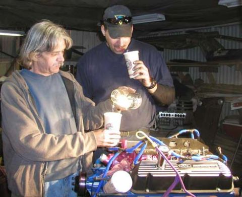1988 Ford Bronco - Two men stand in a metal shop building pouring a drink into paper cups behind an assembled V8 engine on a stand. The engine has a polished…