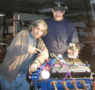 1988 Ford Bronco - Two men pose in a shop behind a freshly assembled V8 engine on a stand, one pointing down at it and the other holding a clear bottle. The b…
