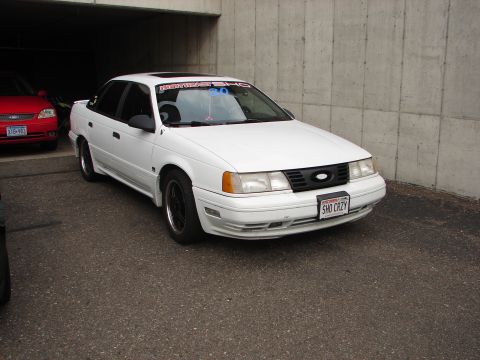1992 Ford Taurus - A white 1992 Ford Taurus is parked under a concrete overhang, with a red car visible to the left.