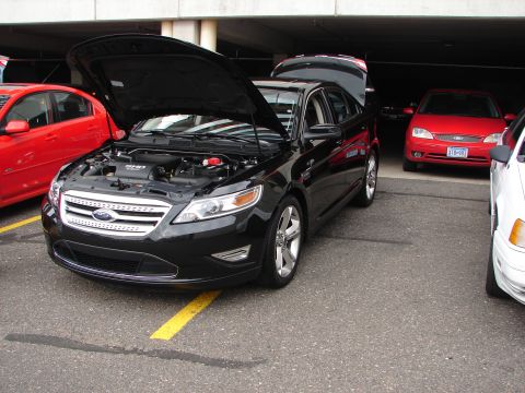 1992 Ford Taurus - A black Ford Taurus is displayed with its hood and trunk open, alongside other vehicles.