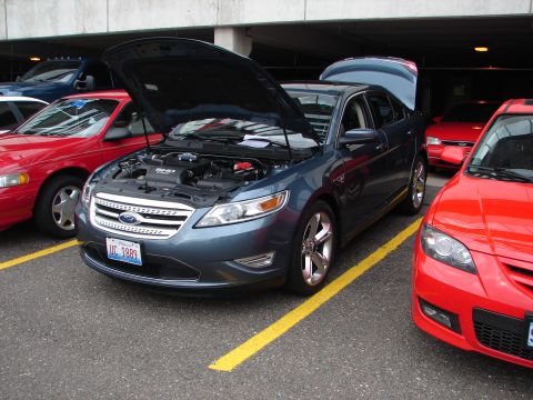 1992 Ford Taurus - A dark blue sedan with both hoods open is parked between two red cars.