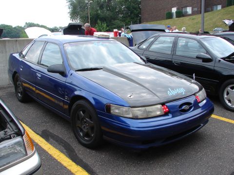 1992 Ford Taurus - A blue 1992 Ford Taurus with a custom hood is parked next to other vehicles at an outdoor gathering.