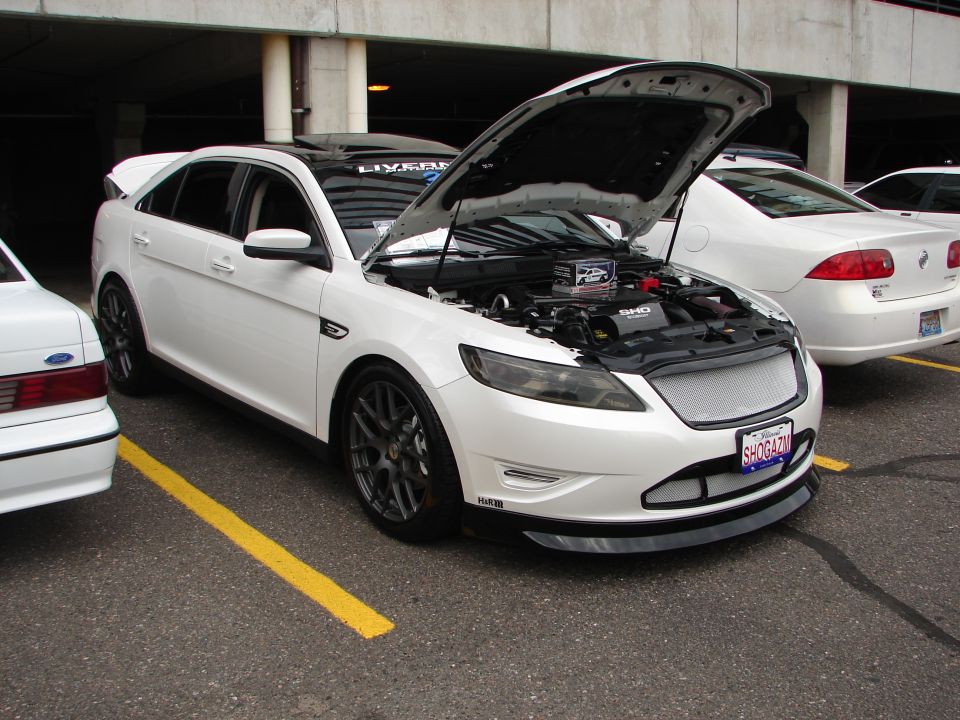 White sedan with hood open, showing engine. Another white car is parked to the left, and a third white car is behind it