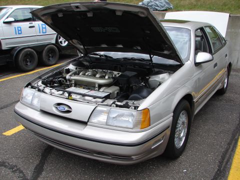 1992 Ford Taurus - A silver 1992 Ford Taurus SHO is displayed with its hood open, revealing the engine. A trailer with racing numbers is visible behind it.