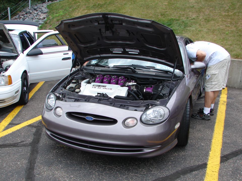 Gray Ford Taurus with hood open, showing V8 32 valve engine. Person looking into engine bay. Another white car next to it