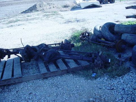 1992 Ford Bronco - Discarded vehicle axles and tires are piled up outdoors on a gravel and grassy area.