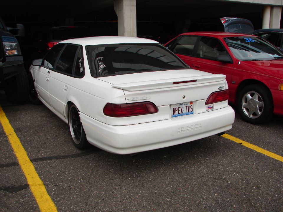 White 1992 Ford Taurus with a spoiler and custom license plate 'APEX THS'. Red car parked to the right