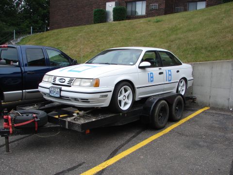 1992 Ford Taurus - A white 1992 Ford Taurus is loaded onto a trailer, with blue racing numbers on its body.