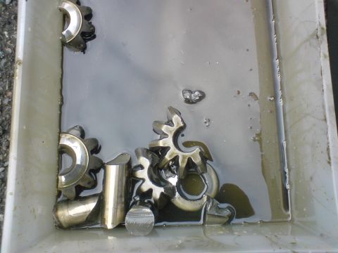 1989 Ford Bronco - Several metal gears are submerged in a shallow pan of dark oil, showing signs of wear and damage.