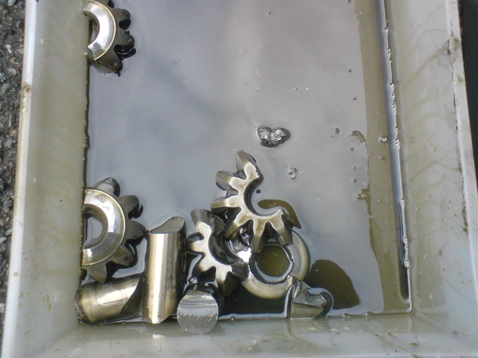metal gears soaking in oil inside a pan, with a 'Supermotors.net' watermark