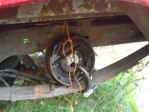1989 Ford Bronco - The rear drum brake assembly of a red vehicle is hanging by a rope, revealing extensive rust on the undercarriage.