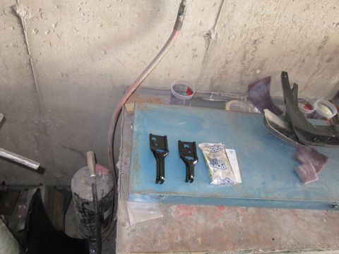 1991 Ford LTD Crown Victoria - Automotive parts, including two black brackets and a fender piece, are laid out on a workbench with a packet of candy.