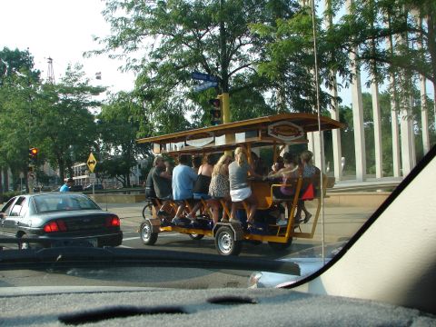 1992 Ford Taurus - A group of people ride a pedal-powered vehicle down a street, passing a car.