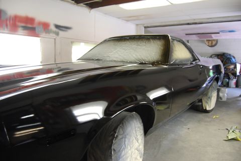 1975 Ford Ranchero - A black coupe is being worked on in a garage, with its windows taped off for painting.