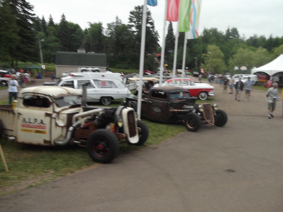 Two hot rod trucks parked on grass and pavement, one white with red lettering, the other rusty brown