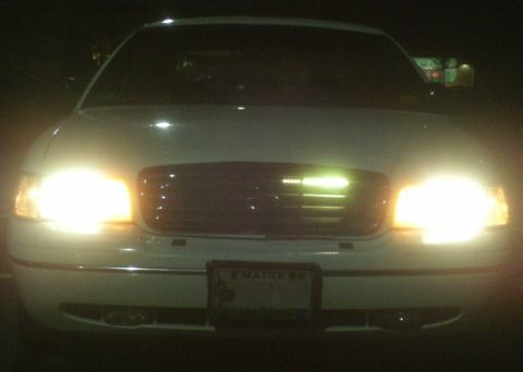 1998 Ford Crown Victoria - A white 1998 Ford Crown Victoria is seen from the front at night with its headlights illuminated.