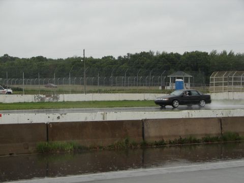 1992 Ford Taurus - A dark sedan drives on a wet track under an overcast sky, with another vehicle visible in the background.