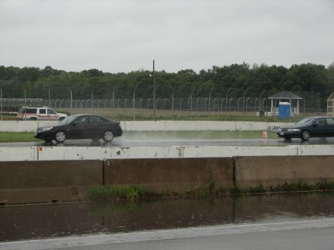 1992 Ford Taurus - Two cars drive on a wet track under an overcast sky, with a white utility vehicle visible in the distance.