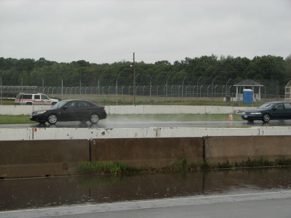 Dark sedan driving on a wet track, with a white utility vehicle in the background