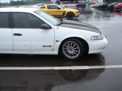 1992 Ford Taurus - A white 1992 Ford Taurus is parked on a wet track, aftermarket wheels and a dark hood.