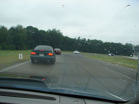 1992 Ford Taurus - A dark 1992 Ford Taurus drives on a wet track with other vehicles in the distance.