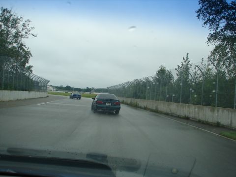 1992 Ford Taurus - Two cars are driving on a wet track under an overcast sky, with safety fencing lining the course.