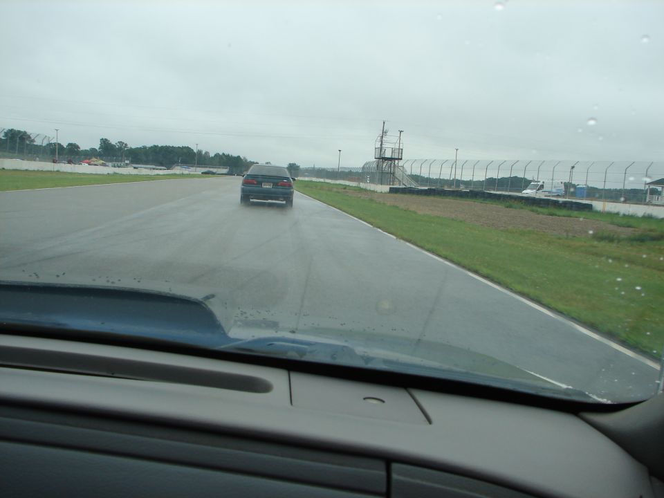Dark sedan driving on a wet racetrack with a guardrail and tower in the background