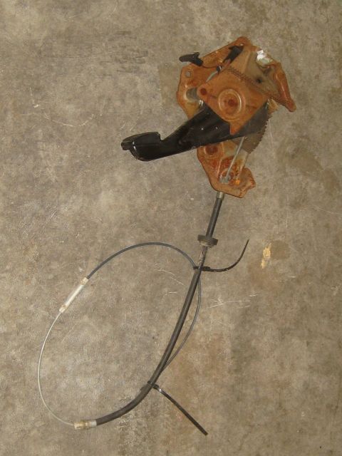 1996 Ford Bronco - A rusty brake pedal assembly with a black pedal pad and cable is shown against a concrete background.