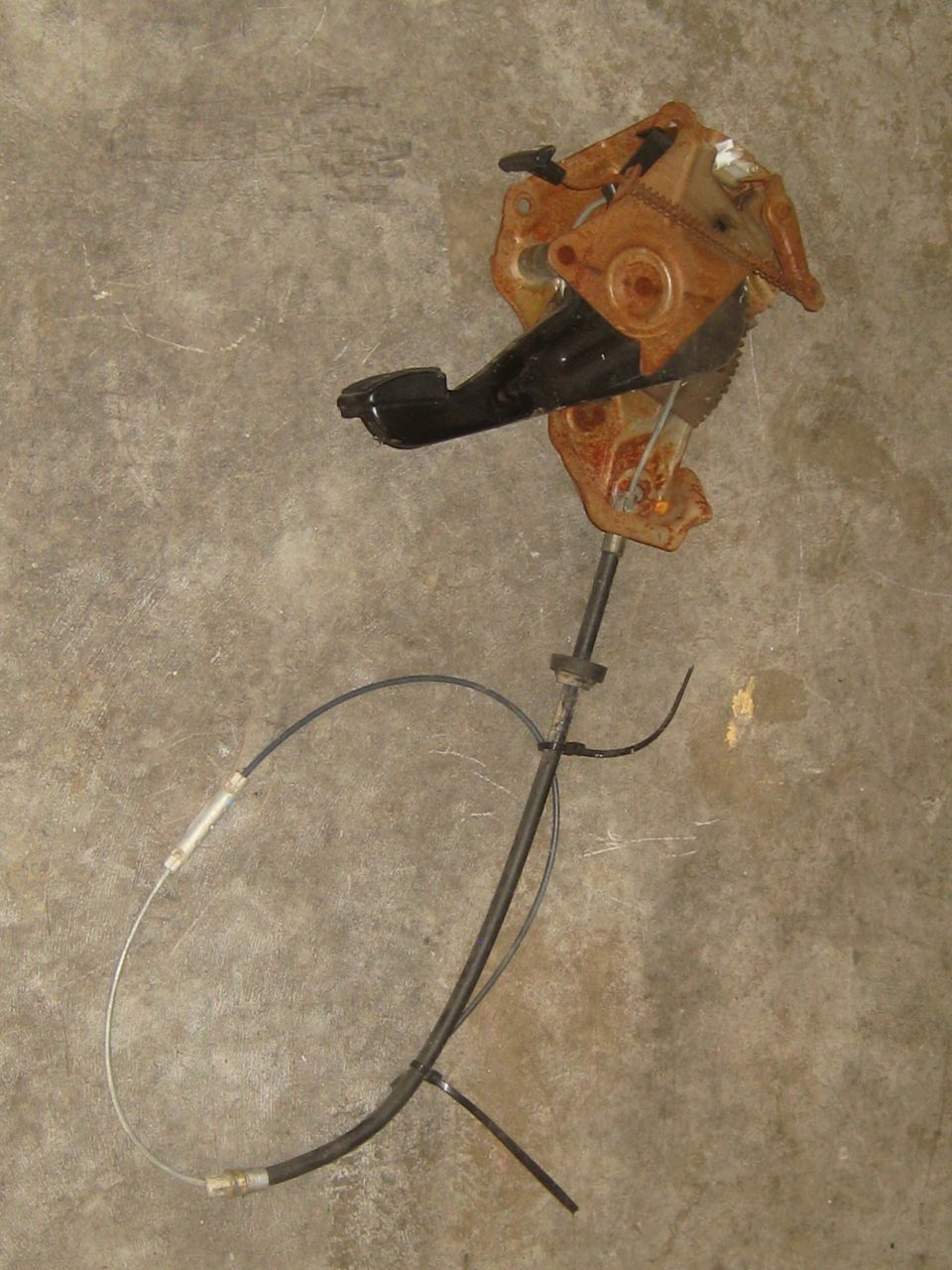 Rusty brake pedal assembly with black pedal pad and attached cable on concrete
