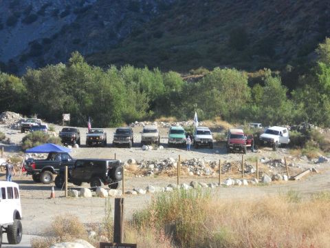 1986 Ford Bronco - A collection of off-road vehicles are parked in a lot, with a mountainous backdrop and trees.