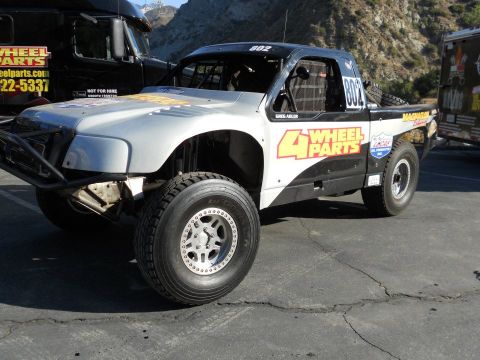 1986 Ford Bronco - This off-road race truck, sponsored by 4 Wheel Parts, is parked next to a large black semi-truck.
