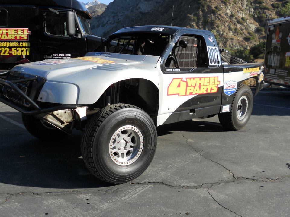 Off-road race truck with "4 Wheel Parts" livery, parked next to a black semi-truck