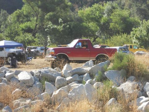 1986 Ford Bronco - A red Ford Bronco is parked on a rocky area with other vehicles in the background.