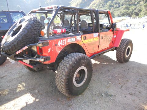 1986 Ford Bronco - Modified red Jeep Wrangler with large off-road tires and a spare tire carrier at the rear. A dark vehicle is to the left.