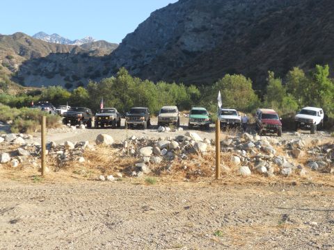 1986 Ford Bronco - A group of off-road vehicles are parked in a line on a dirt path, with mountains and trees behind them.