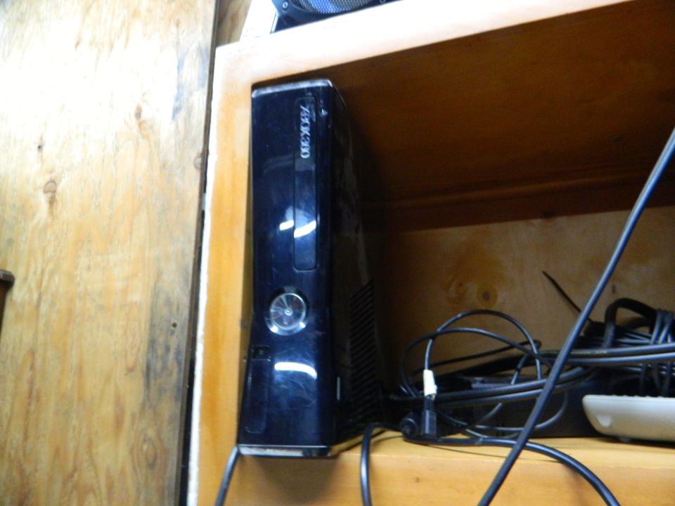 Black Xbox 360 console standing vertically on a wooden shelf, with cables behind it