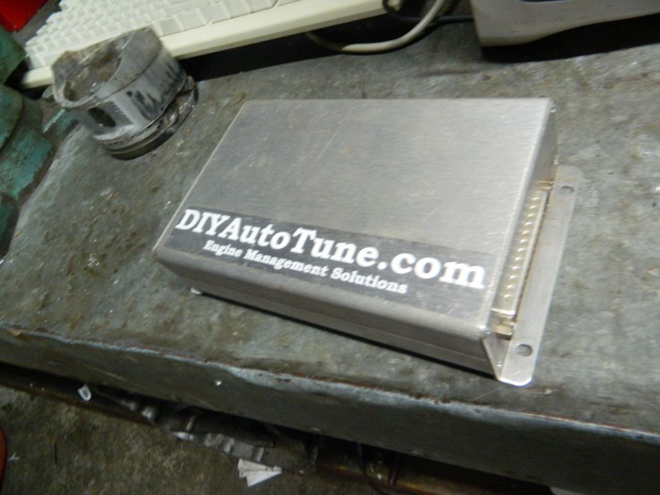Silver engine control unit with DIYAutoTune.com sticker and a piston in the background