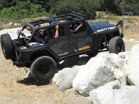 1986 Ford Bronco - A black Jeep Wrangler is shown off-roading over large rocks, with "Smittybilt" and "Pro Comp" branding visible.