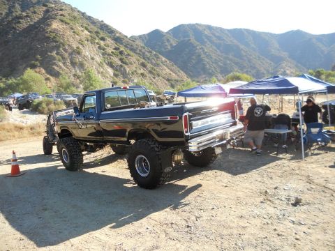 1986 Ford Bronco - A lifted black Ford F-Series truck from the mid-1970s is parked outdoors with other vehicles and tents in the background.