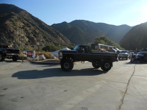 1986 Ford Bronco - A lifted dark blue pickup truck is parked outdoors with mountains in the background. Other vehicles and people are visible.