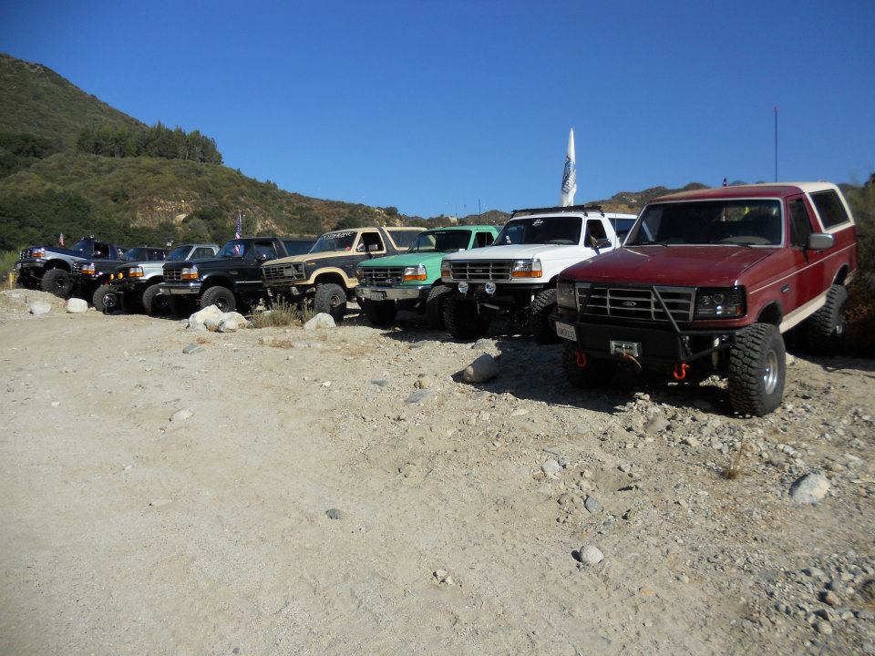 Several Ford Broncos parked on a dirt lot with hills in the background. The red Bronco in front has a custom bumper