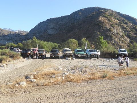 1986 Ford Bronco - A group of off-road vehicles are gathered on a dirt path, with a large mountain range visible behind them.
