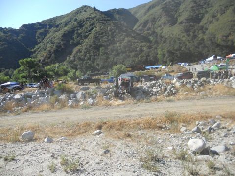 1986 Ford Bronco - Multiple off-road vehicles are parked and driving in a , mountainous terrain with tents set up.