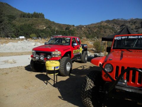 1986 Ford Bronco - Two off-road vehicles, a red GMC pickup and an orange Jeep, are parked in a dry, mountainous area under a clear blue sky.