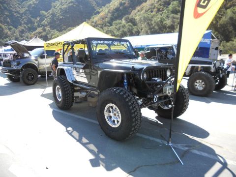 1986 Ford Bronco - A modified black Jeep Wrangler is displayed at an outdoor event, with other vehicles and tents in the background.