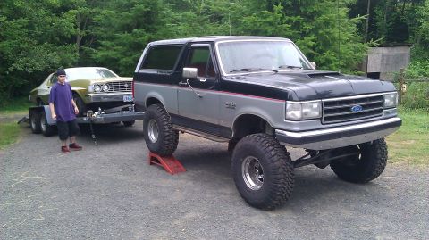 1973 Ford Torino - A lifted Ford Bronco tows a 1975 Ford car on a trailer, with a person standing nearby.