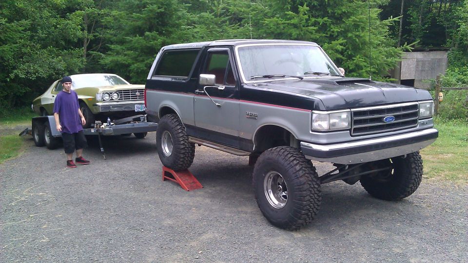 Olive green 1975 Ford car on a trailer, towed by a lifted black and gray Ford Bronco