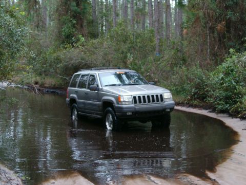 1991 Ford Bronco - A silver Jeep Grand Cherokee navigates a water crossing in a wooded area.