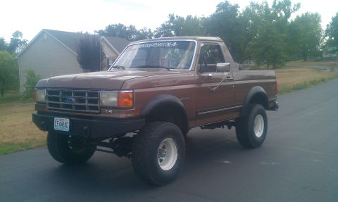 1987 Ford Bronco - This lifted 1987 Ford Bronco features a brown exterior and white rims, parked on a paved surface with houses in the background.
