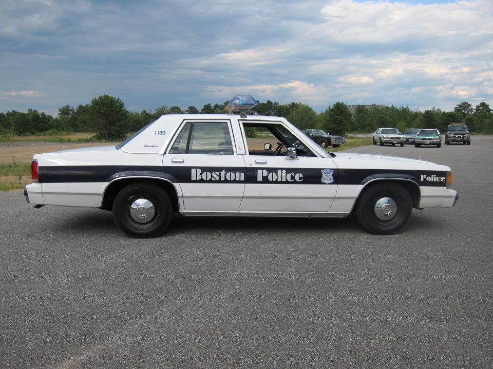 Side view of a white Boston Police car with a dark stripe and "Boston Police" text. Other cars are parked in the background